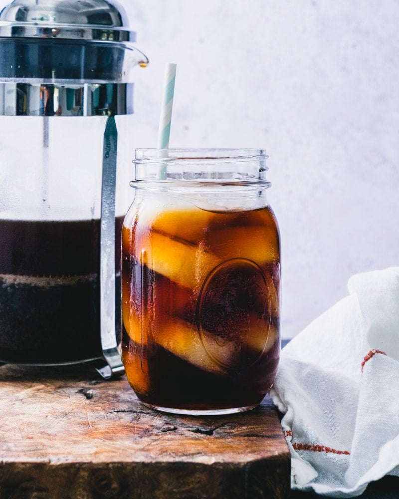 A French press filled with ground coffee sitting next to a mason jar glass with coffee over ice and a straw.