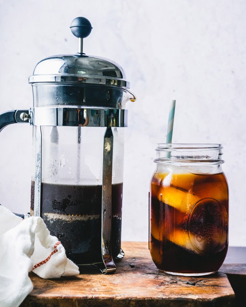 A French press filled with ground coffee sitting next to a mason jar glass with coffee over ice and a straw.