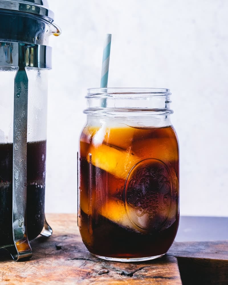 A French press filled with ground coffee sitting next to a mason jar glass with coffee over ice and a straw.
