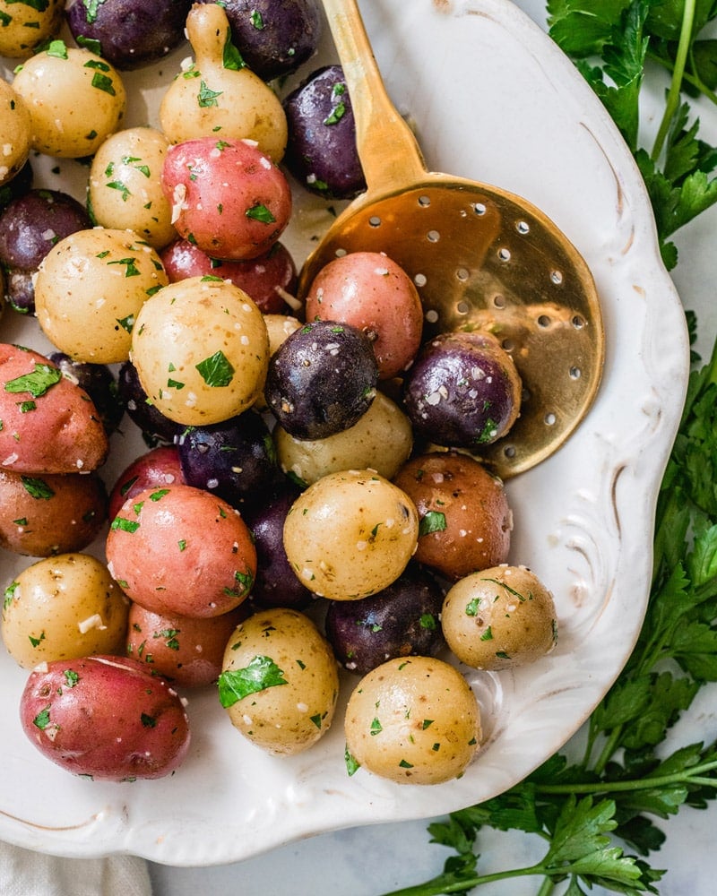 Garlic steamed potatoes, purple, yellow, and red, on a white plate and surrounded by parsley leaves.