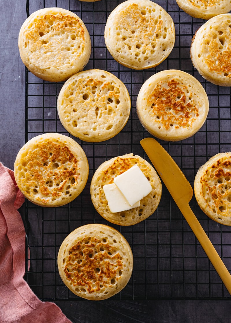Crumpets on a cooling rack with buter slices on one and a butter knife next to it