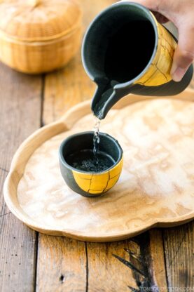 sake being served from a black vessel into a cup on a wooden tray