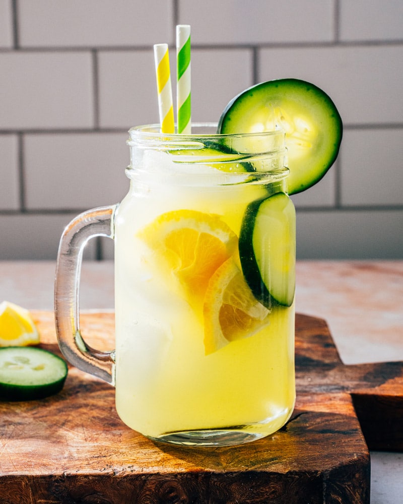 Cucumber Lemonade in a mason jar wtih a handle and two straws, garnished with lemons and cucumbers