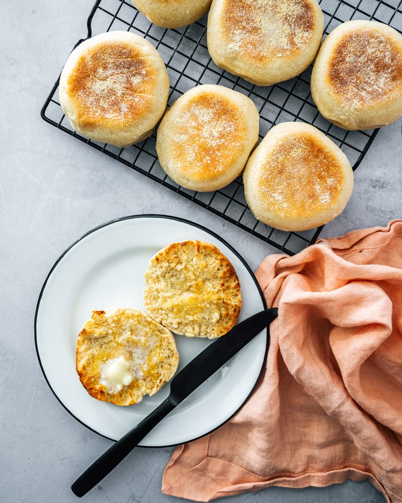 English muffin sittin on a cooling rack, and one cut in half and covered in butter on a plate