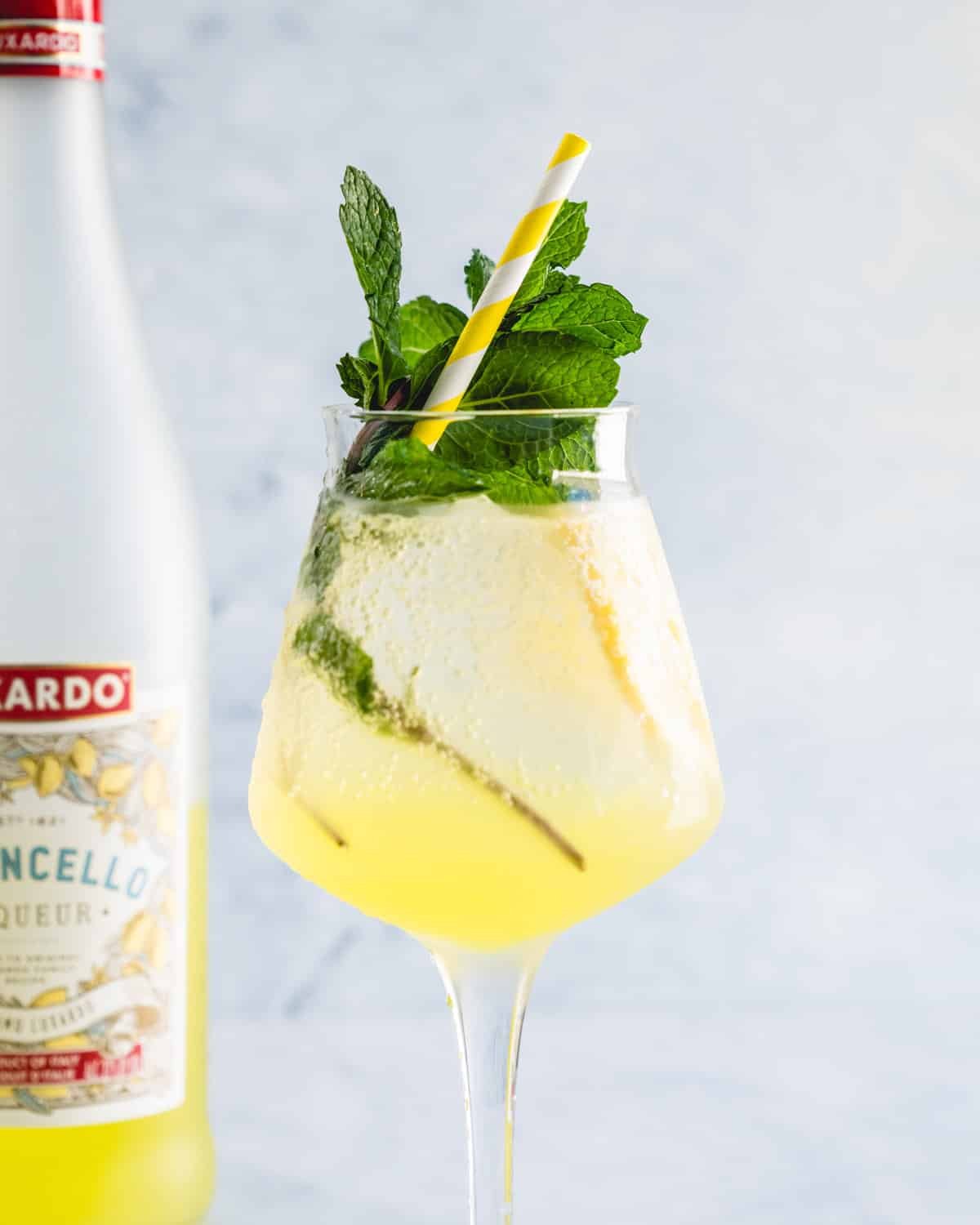 Limoncello spritz in a stemmed glass with mint and a straw next to a bottle of Limoncello