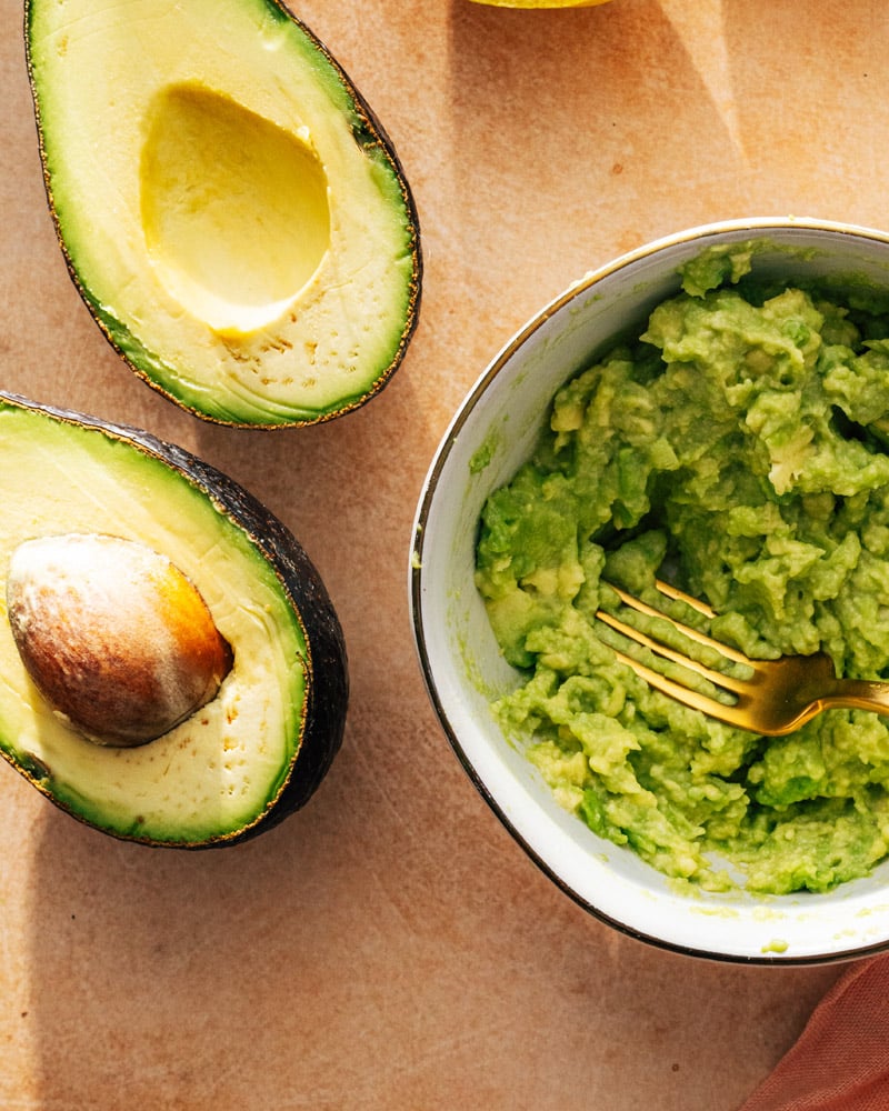 Avocado cut open alongside a bowl of mashed avocado.