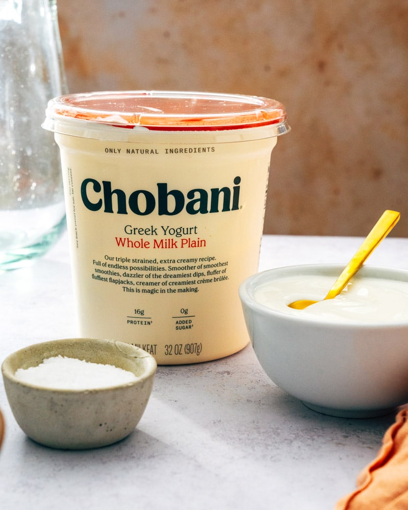 A tub of chobani greek yogurt sitting on a table with a small bowl of sugar, and a glass bowl of yogurt