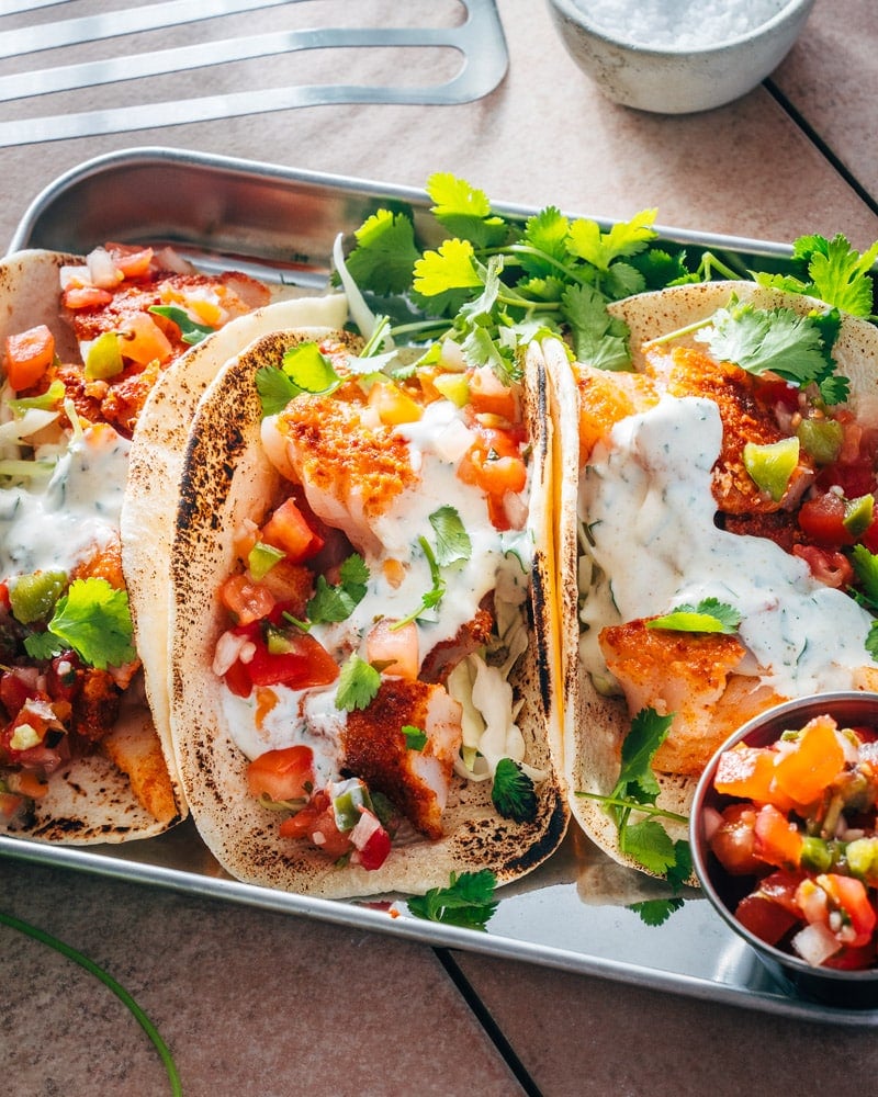 Three cod fish tacos on a tray with a small bowl of pico de gallo next to them and cilantra spread over the top