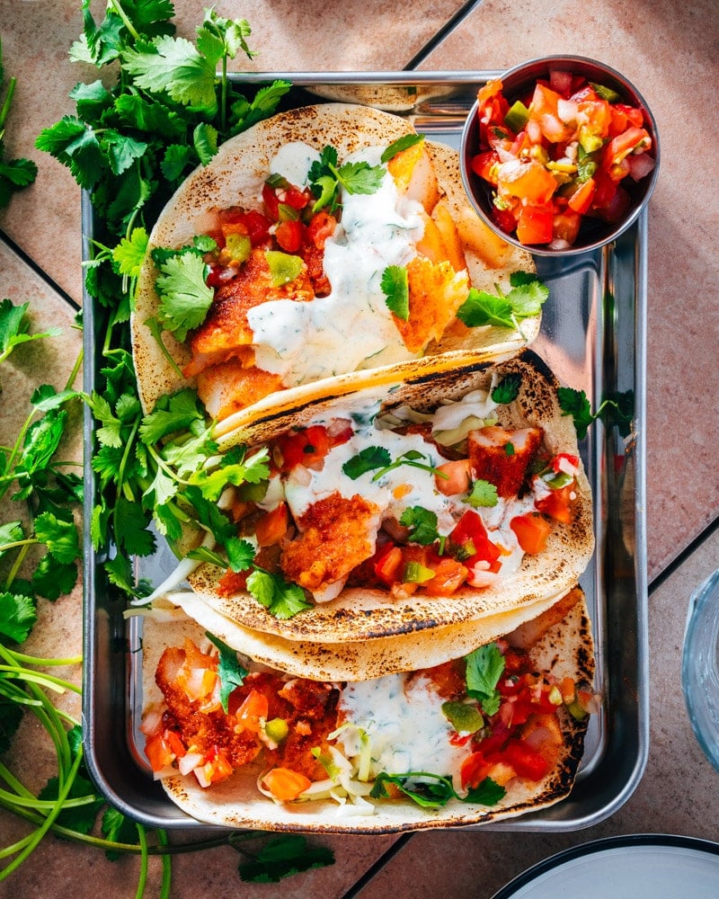 Three cod fish tacos on a tray with a small bowl of pico de gallo next to them and cilantra spread over the top
