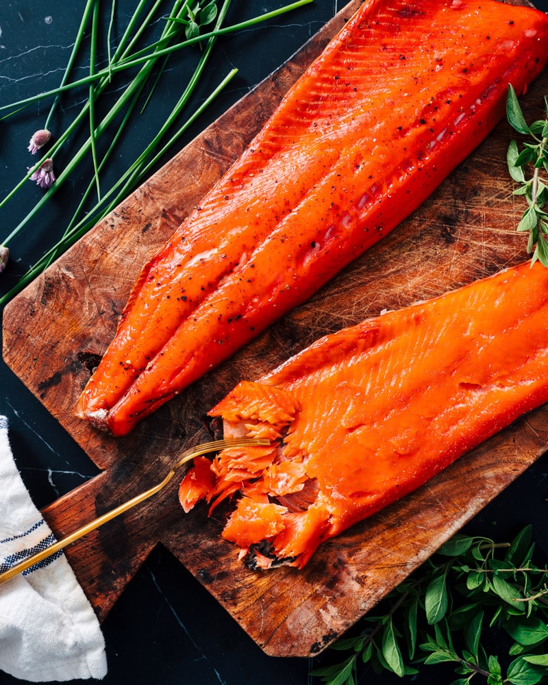 Two smoked salmon fillets on a cutting board, with one being shredded by a fork