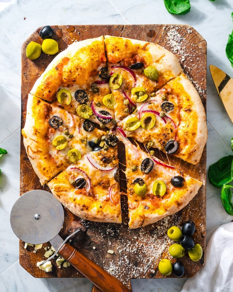 Olive Pizza on a cutting board with red onion, capers, and green and black olives with a pizza cutter sitting next to it