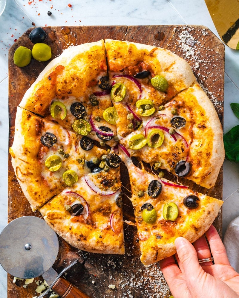 Olive Pizza on a cutting board with red onion, capers, and green and black olives with a hand grabbing a slice
