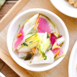 A small white bowl filled with vibrant Japanese pickles, including slices of radish, napa cabbage, and carrots, served on a wooden tray.
