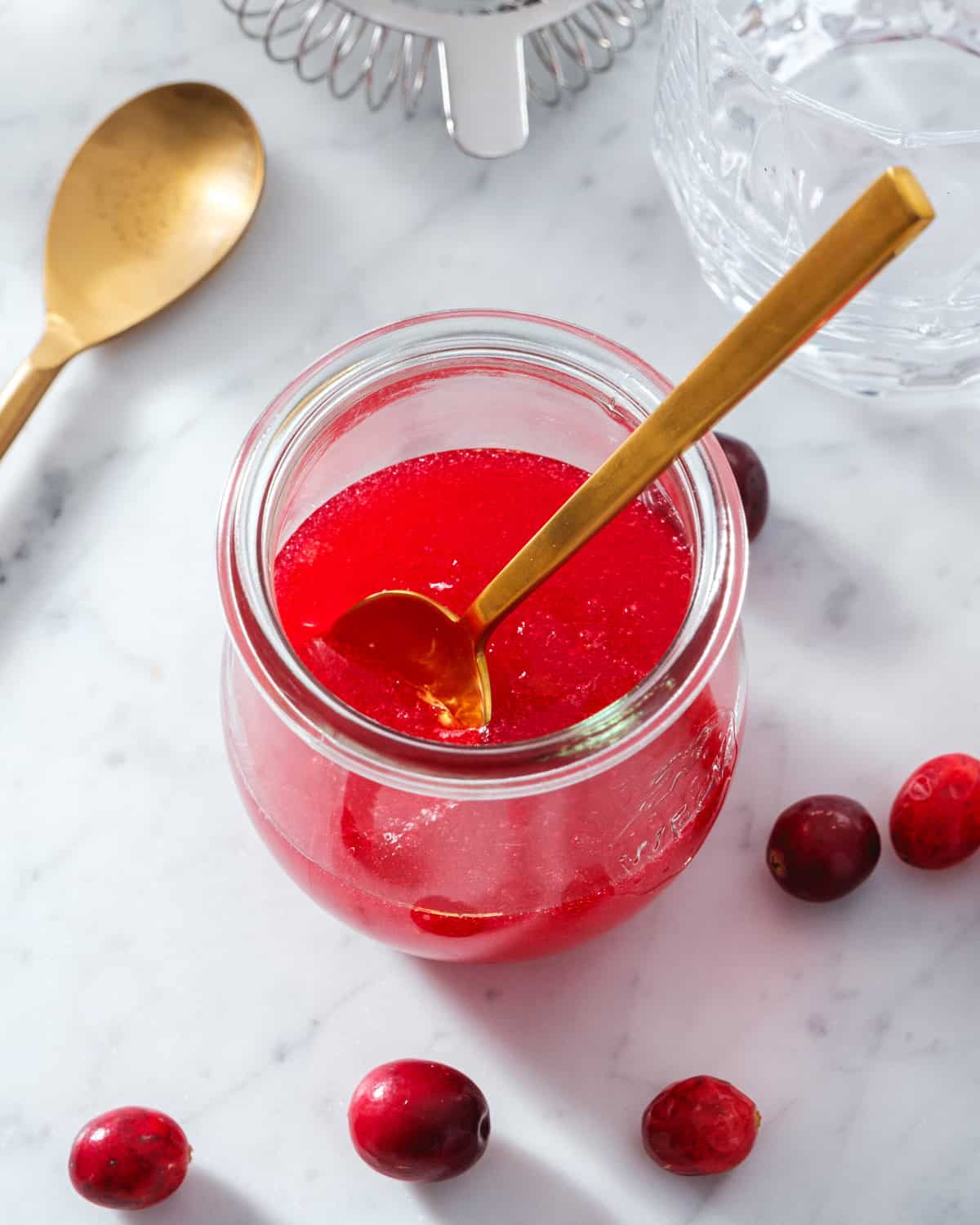 Cranberry Simple Syrup in jar with spoon and cocktail strainer in background
