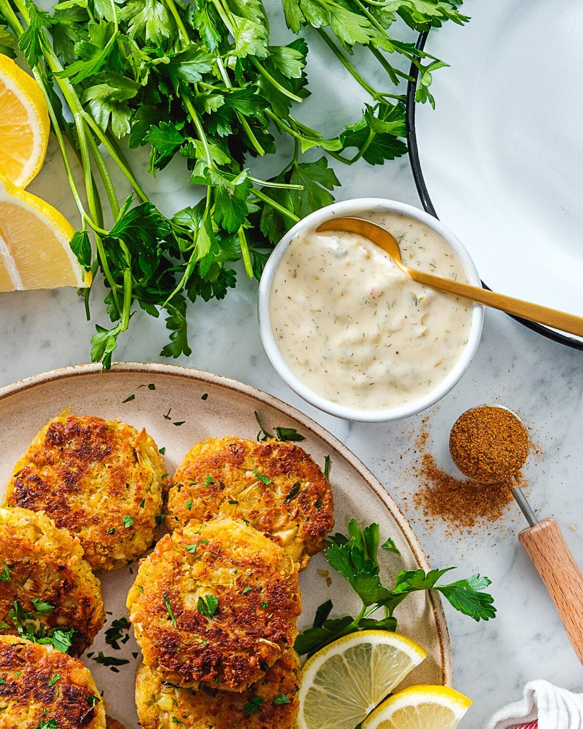 Vegan Crab Cakes on a plate with lemon wedges next to a side bowl of Vegan Tartar Sauce, a tablespoon of seasoning, and green herbs