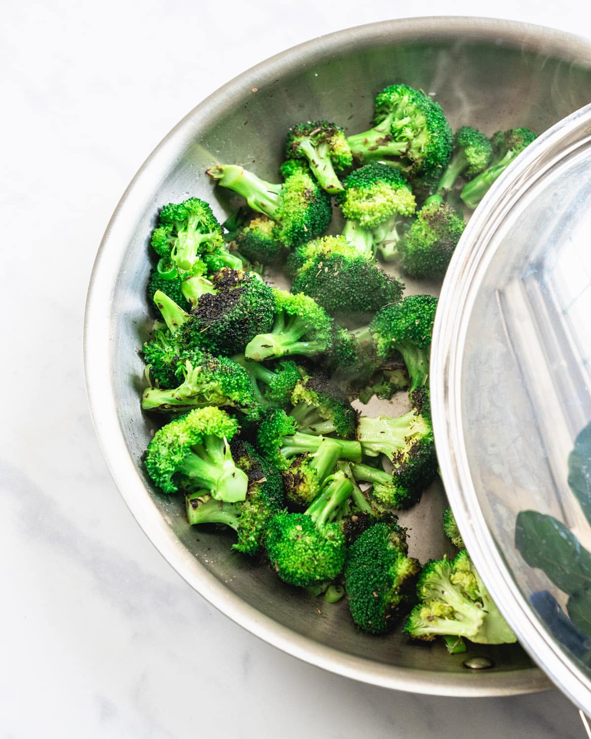 Broccoli in a large skillet with a lid being placed to steam