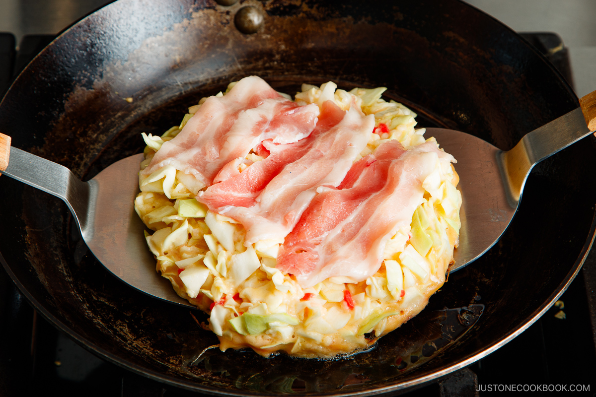 A round okonomiyaki cabbage pancake topped with raw pork slices is being cooked in a black skillet, held by two wide metal spatulas on either side. The okonomiyaki pancake is golden at the edges.