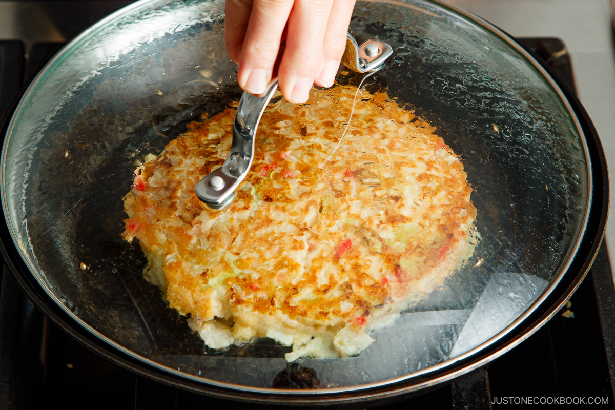 A hand lifts the glass lid from a frying pan, revealing a golden-brown okonomiyaki pancake with crispy edges sizzling on the stovetop.