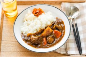 A round white plate containing Japanese beef curry consisting of tender beef, potatoes, carrots, mushrooms, and curry roux, served over steamed rice.