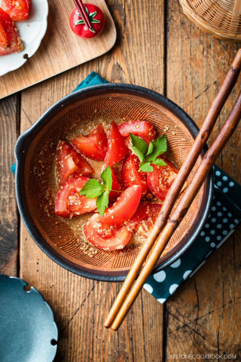 A Japanese black mortar containing cut tomato wedges dressed in ground sesame ponzu seasoning, garnished with mitsuba leaves on top.