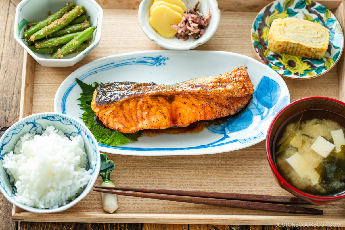 A Japanese meal consisting of small dishes or bowls, each plating Teriyaki Salmon, steamed rice, miso soup, pickles, tamagoyaki, and green bean gomaae.