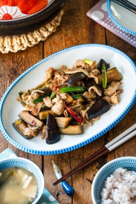 An oval plate with scalloped edge containing Pork and Eggplant Stir Fry served with multigrain rice and miso soup.