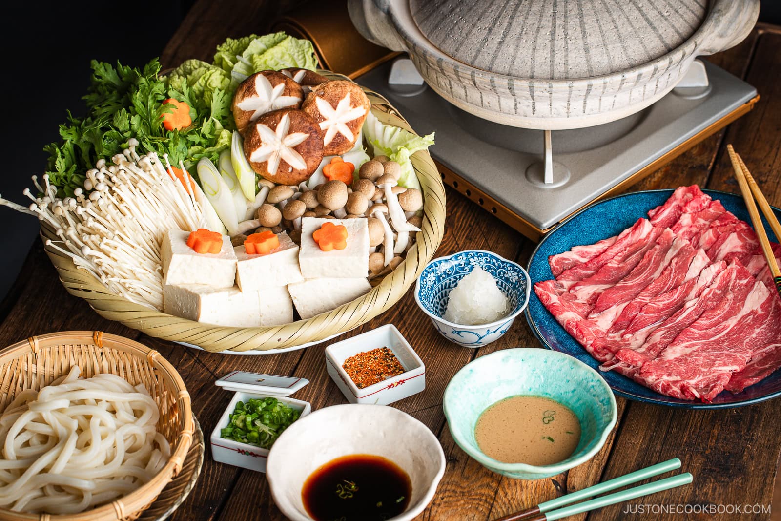 A Japanese shabu-shabu hot pot setup with a donabe pot, thinly sliced beef, tofu, assorted mushrooms, vegetables, udon noodles, and dipping sauces arranged on a wooden table.