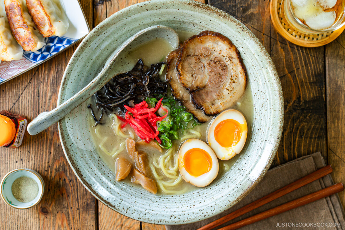 A rustic bowl containing noodles with tonkotsu broth, topped with chashu, ramen eggs, menma, kikurage, ginger, and chopped green onion. On the table, a plate of crispy gyoza and a glass of mugicha are served together.