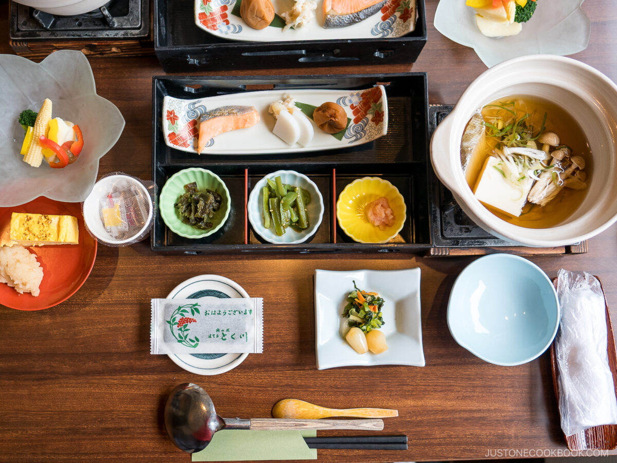 A traditional Japanese breakfast set with grilled fish, tofu hot pot, pickled vegetables, tamagoyaki, rice, miso soup, and small side dishes, neatly arranged on a wooden table.