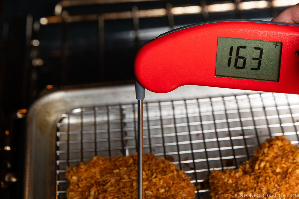 A red digital meat thermometer reads 163&deg;F while inserted into a breaded piece of meat on a wire rack over a baking tray inside an oven.