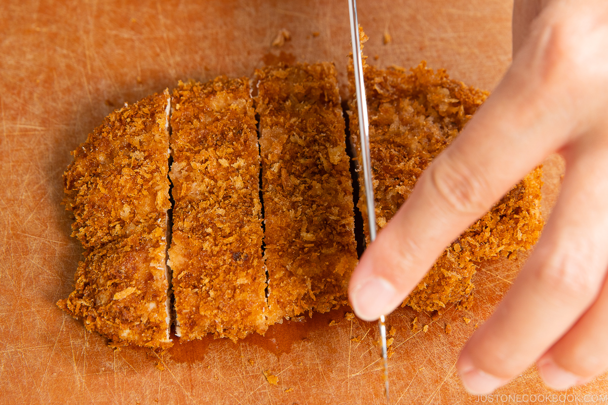A hand slicing a breaded, golden-brown fried cutlet on a wooden cutting board with a sharp knife.