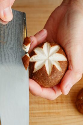 A person uses a knife to carve a decorative star pattern into the cap of a shiitake mushroom on a wooden cutting board.