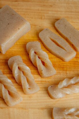 Rectangular and twisted pieces of beige konnyaku (yam cake) are arranged on a wooden cutting board. The pieces vary in shape, with some flat and others braided.