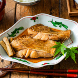 Two pieces of glazed simmered fish are served in a decorative plate with sauce, garnished with burdock root and a green maple leaf, with chopsticks on the side, on a rustic wooden table.