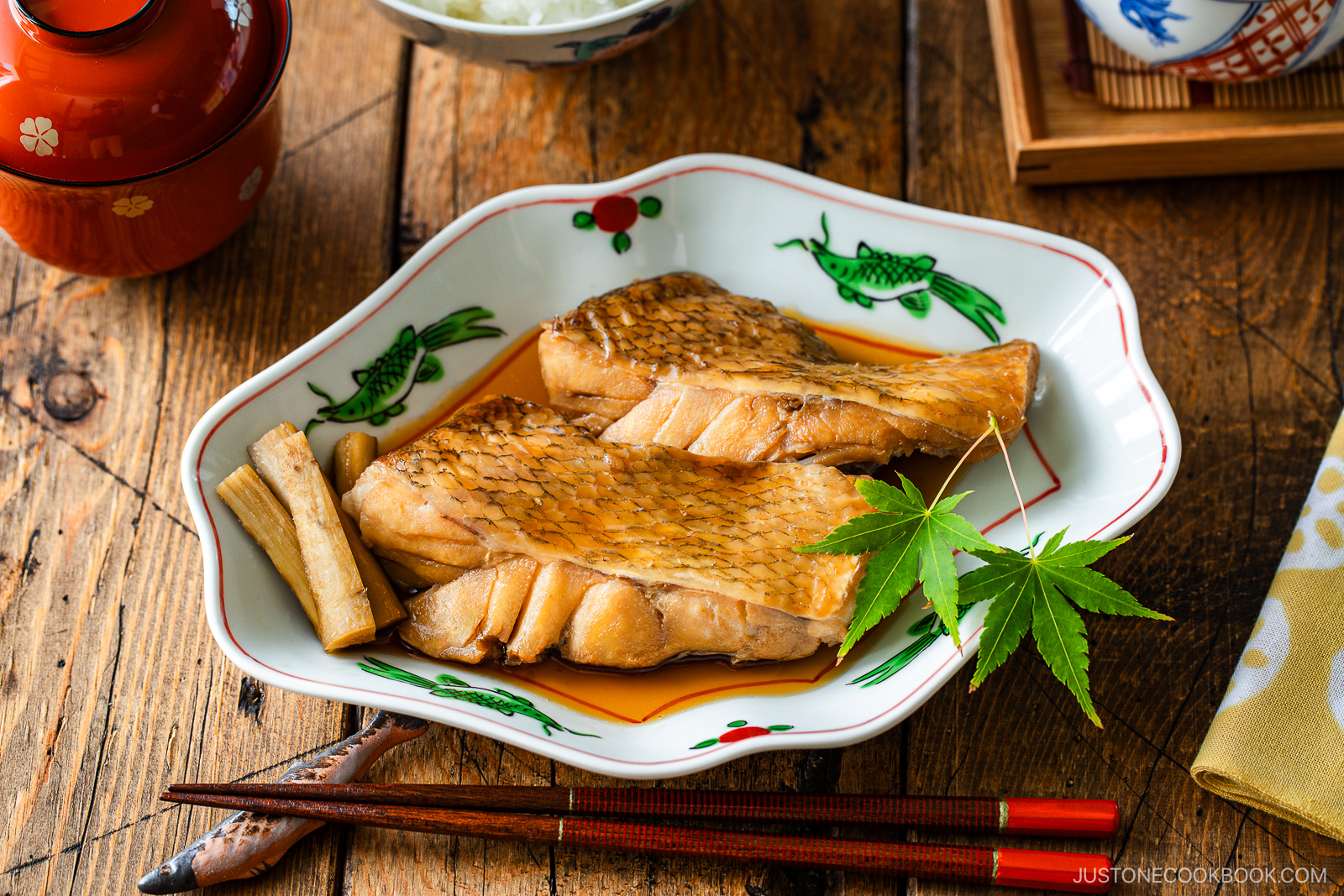 Two pieces of glazed simmered fish are served in a decorative plate with sauce, garnished with burdock root and a green maple leaf, with chopsticks on the side, on a rustic wooden table.