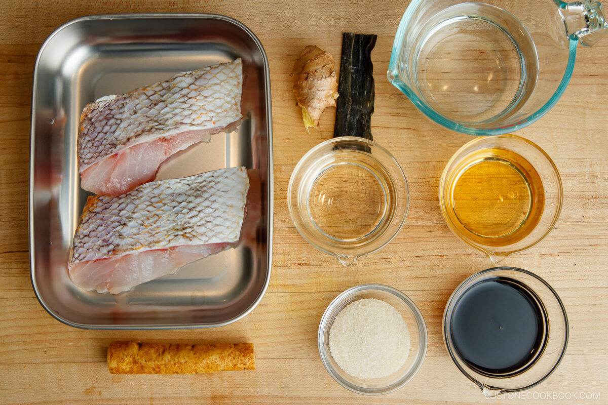 Raw fish fillets in a metal tray next to ginger, kombu seaweed, water in a measuring cup, mirin, sake, soy sauce, sugar, and a stick of yaki-fu on a wooden surface.