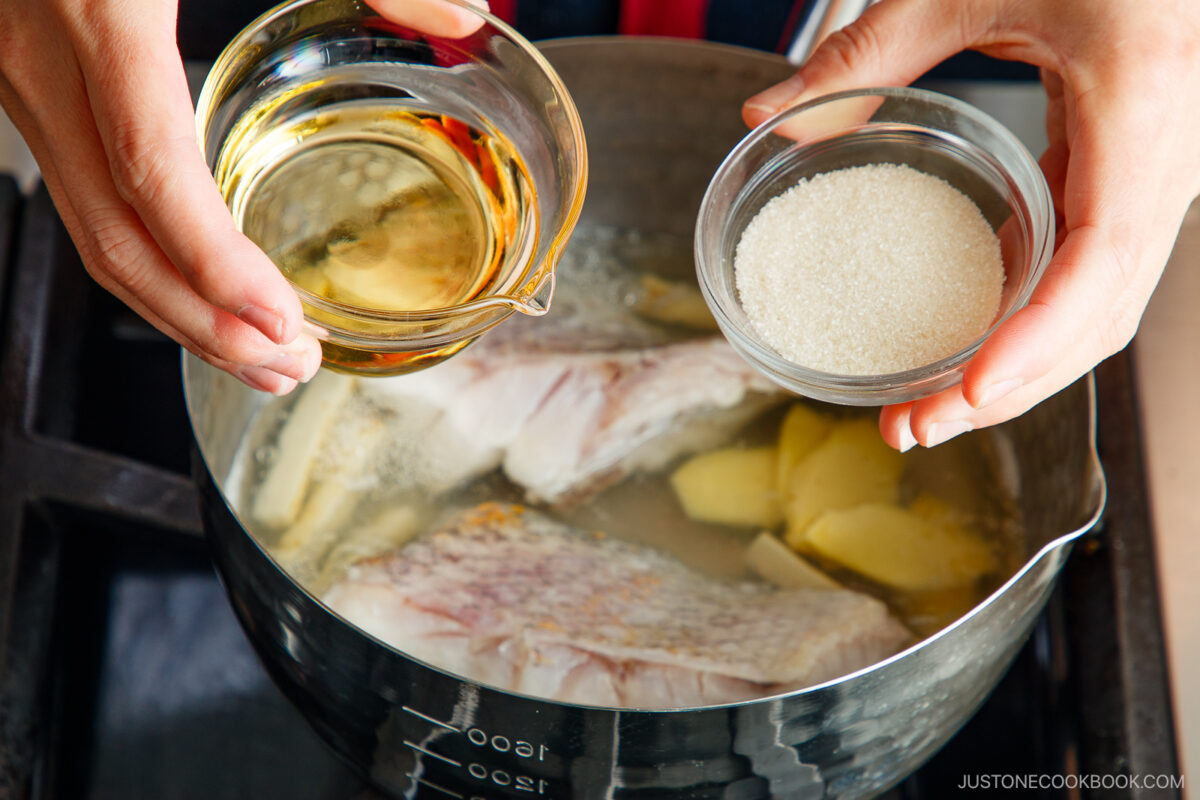 A person holds a small bowl of sugar and a small bowl of liquid over a pot with fish fillets and ginger slices simmering in water on a stovetop.
