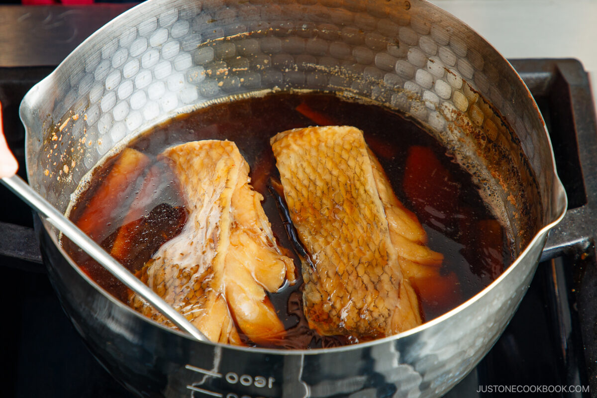 Two pieces of fish simmer in a hammered metal pot filled with a dark, savory broth. A metal spoon is stirring the pot, and the fish skin and texture are visible. The scene is set on a stovetop.