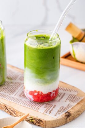 A glass with a clear straw contains a layered drink of red puree at the bottom, white cream in the middle, and green matcha on top, sitting on a wooden board lined with printed paper.