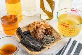 A bamboo tray with dried kombu, shiitake mushrooms, small fish, and bonito flakes sits on a white surface, surrounded by measuring cups and bottles filled with golden and clear broths. A striped cloth is nearby.