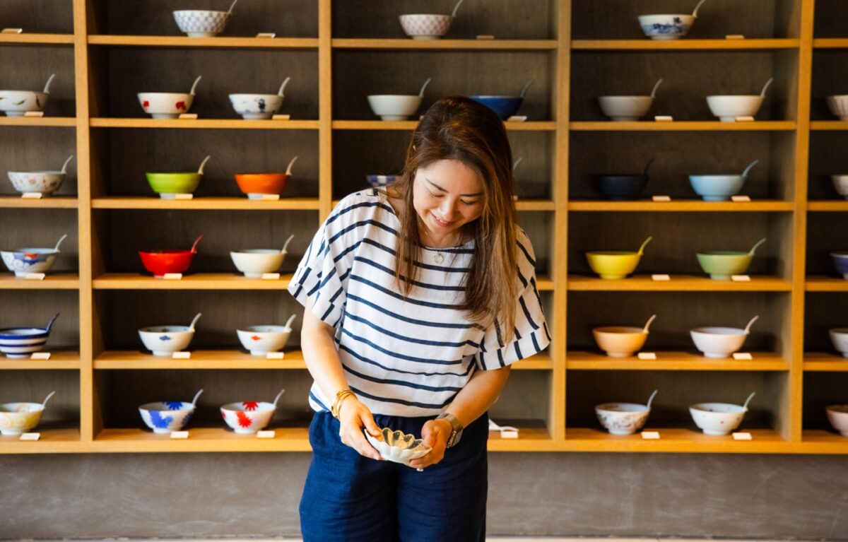 Colorful Japanese rice bowls displayed on a wooden shelf at a ceramics shop, with a smiling woman selecting a bowl, highlighting traditional tableware and cultural craftsmanship.