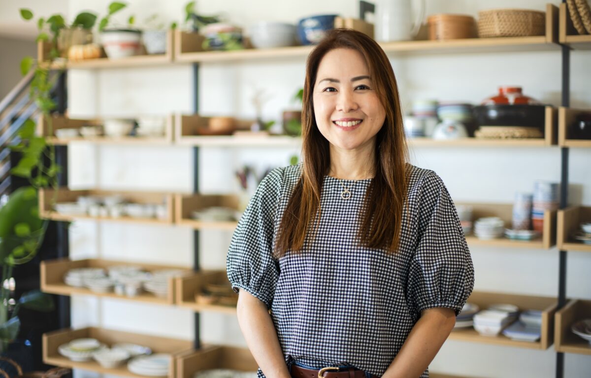A woman with long hair, wearing a checkered blouse, stands smiling in front of shelves filled with various dishes and pottery in a well-lit room&mdash;perfectly capturing the warm atmosphere of About Fancy Foods Show.