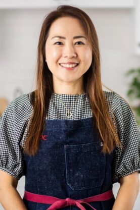 Smiling woman in a kitchen wearing a black and white checkered top and navy apron, showcasing Japanese home cooking and culinary expertise.