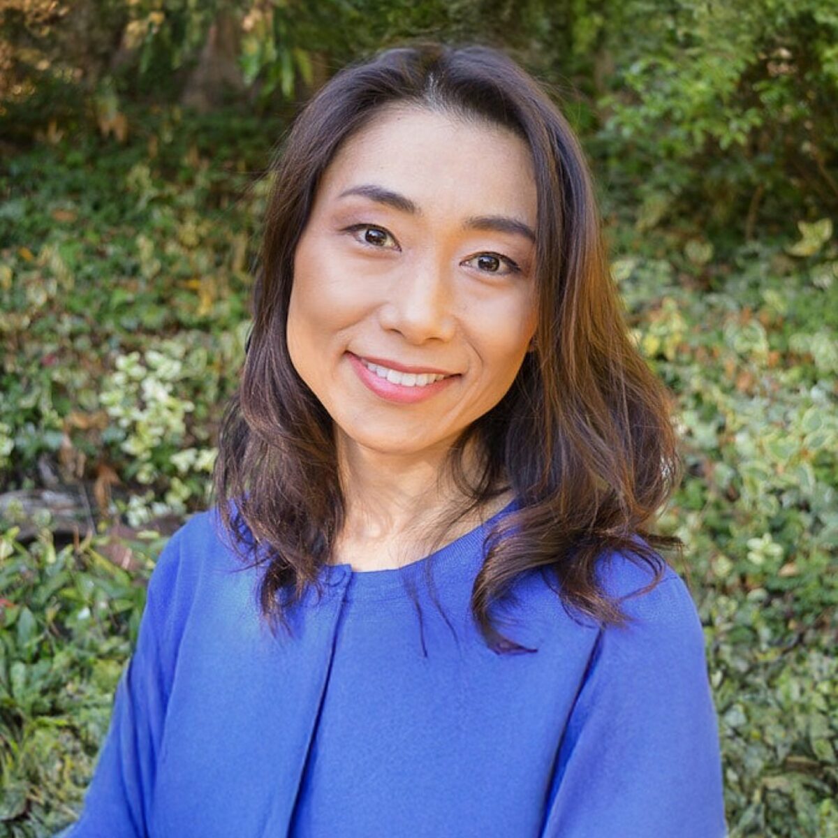Portrait of an Asian woman smiling outdoors, wearing a bright blue top with a lush green background; perfect for highlighting cultural cuisine or lifestyle content on Fancy Foods Show.