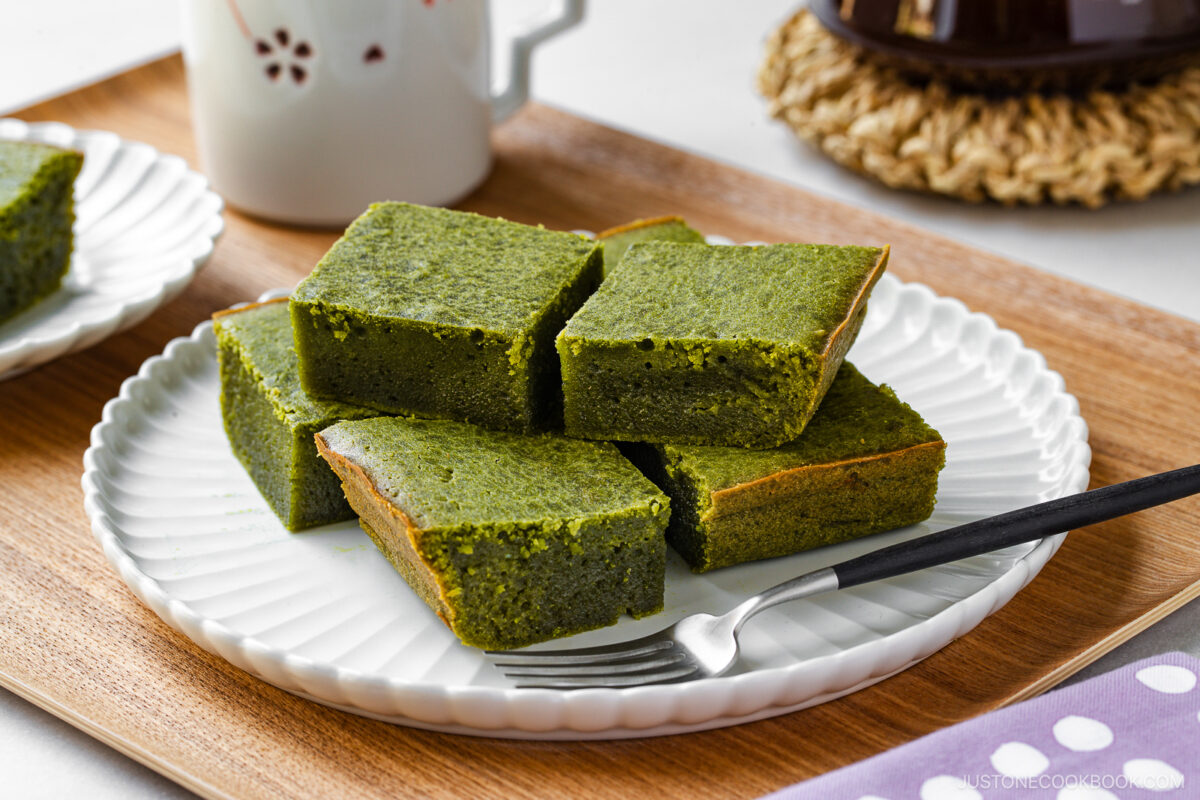 A white plate with six square pieces of green matcha brownies is placed on a wooden tray. A black-handled fork rests beside the brownies, and a white mug sits in the background.