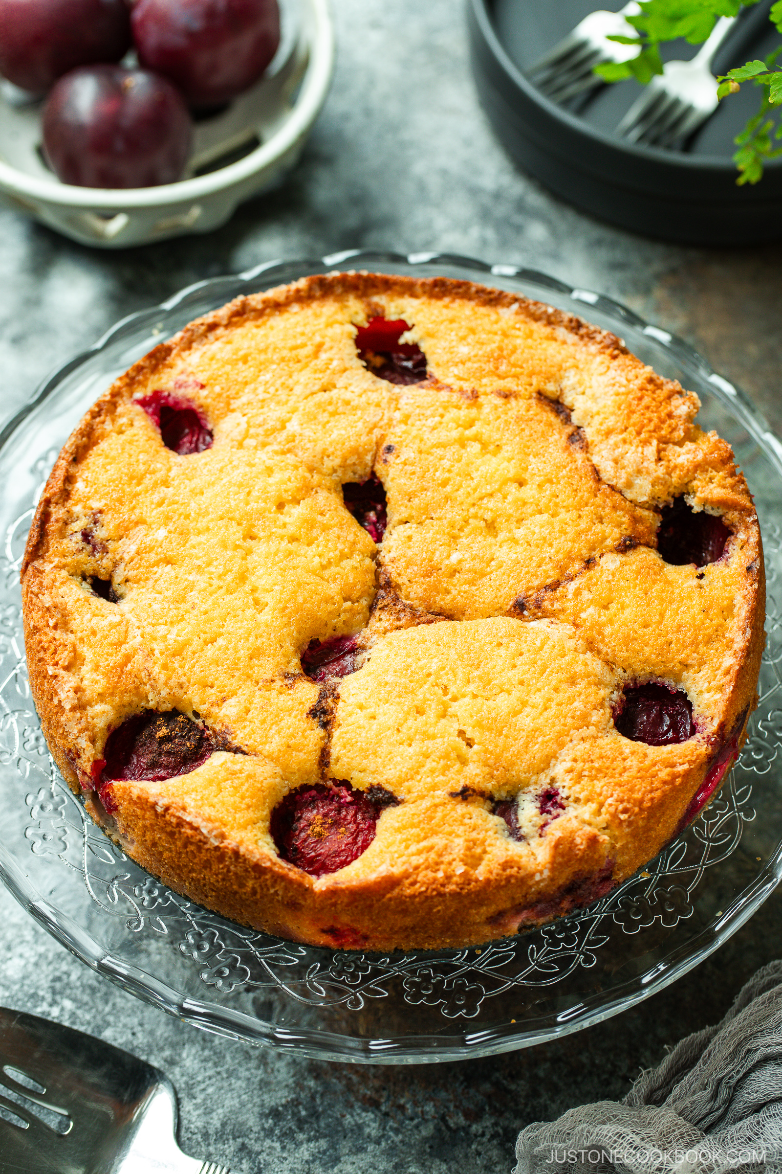 A golden-brown plum cake studded with halved plums sits on a glass cake stand. In the background, whole plums and a black plate with green garnish are visible.