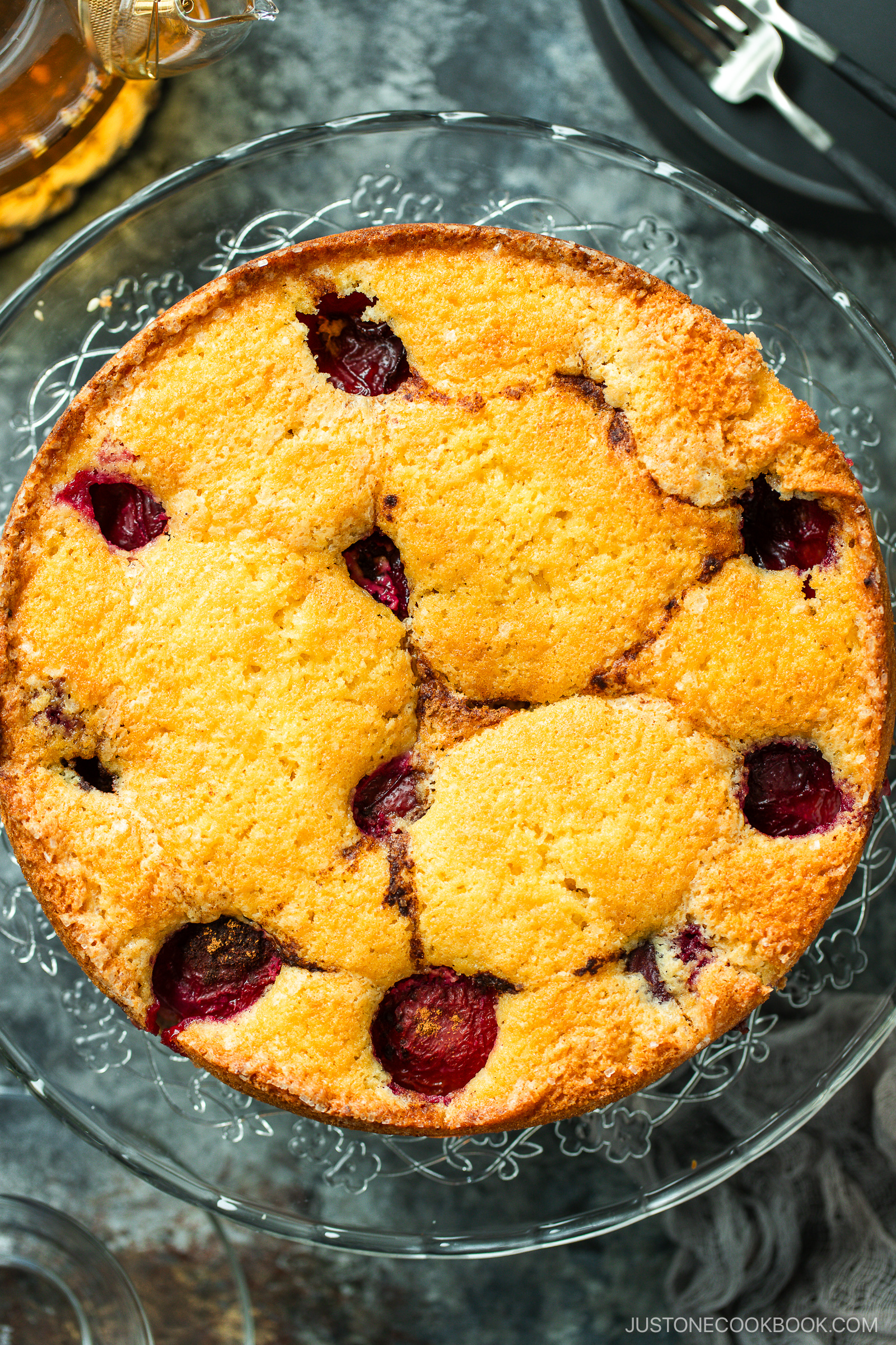 A round golden-brown cake with whole cherries baked inside, displayed on a glass cake stand, viewed from above.
