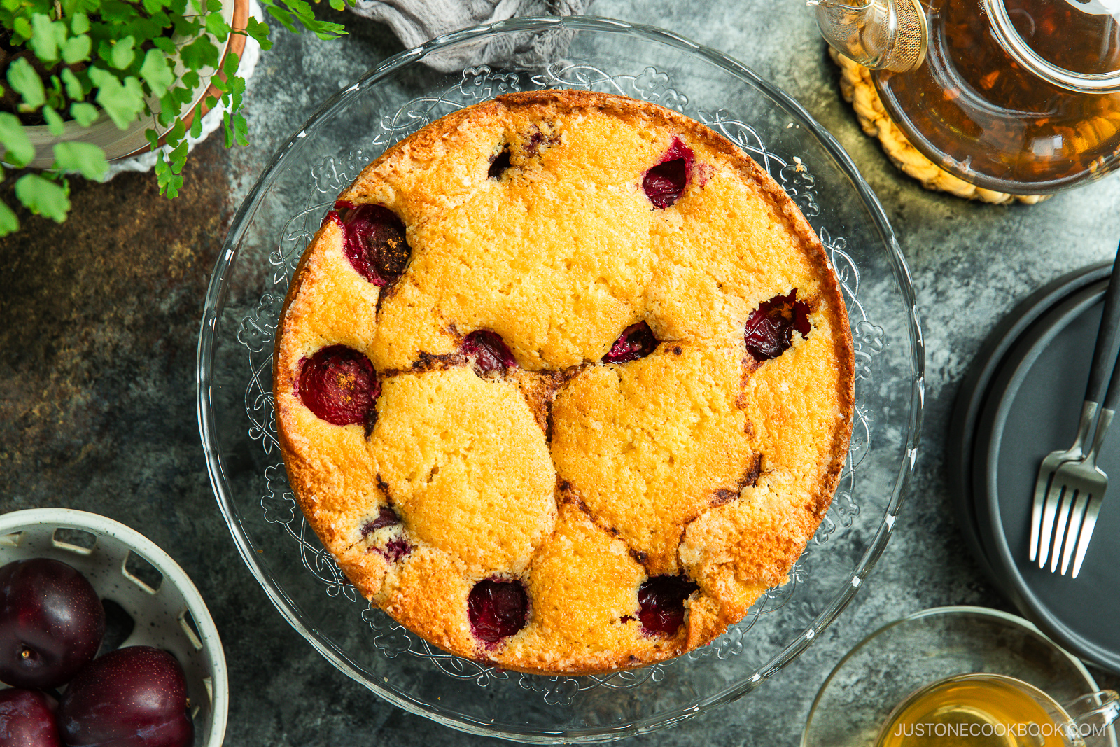A round, golden-brown plum cake with visible plum halves sits on a glass cake stand. Nearby are fresh plums, a teapot, black plates with utensils, and green leafy plants on a textured tabletop.