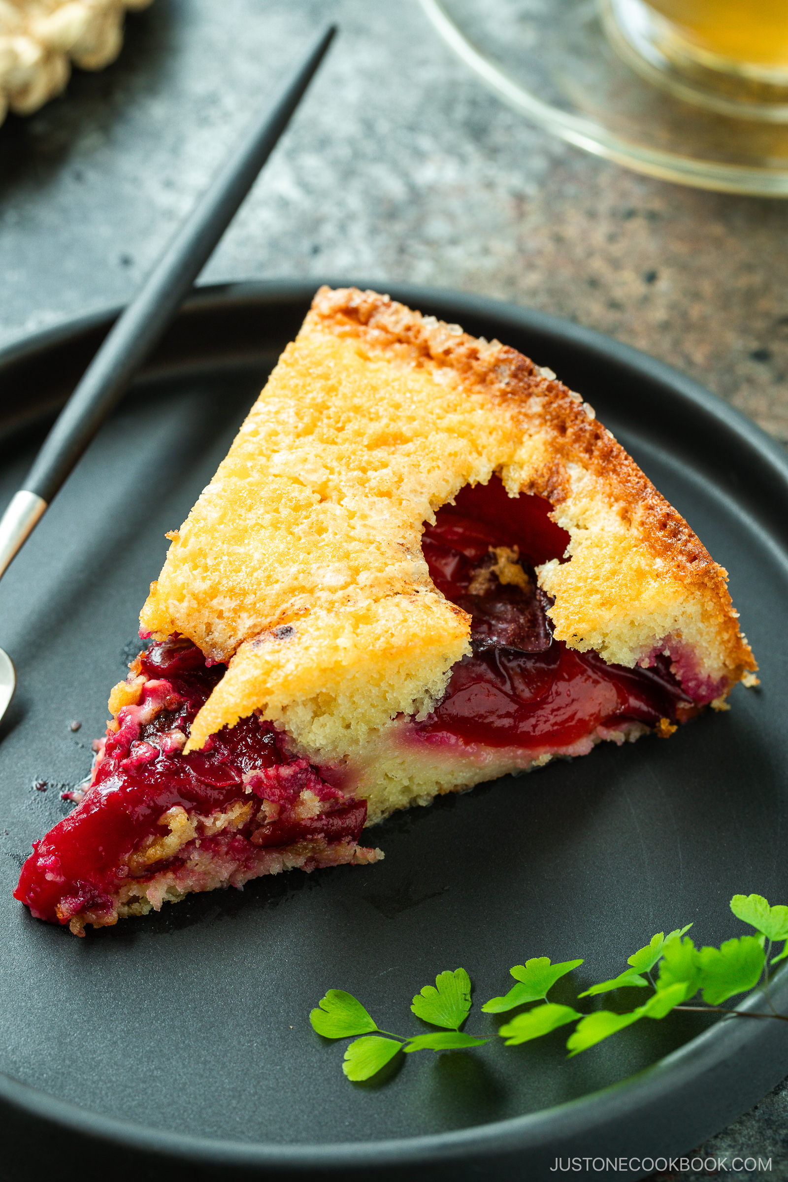A slice of plum cake with a golden, crumbly top sits on a dark plate beside a fork and a small green herb garnish. Juicy baked plums are visible inside the moist cake.