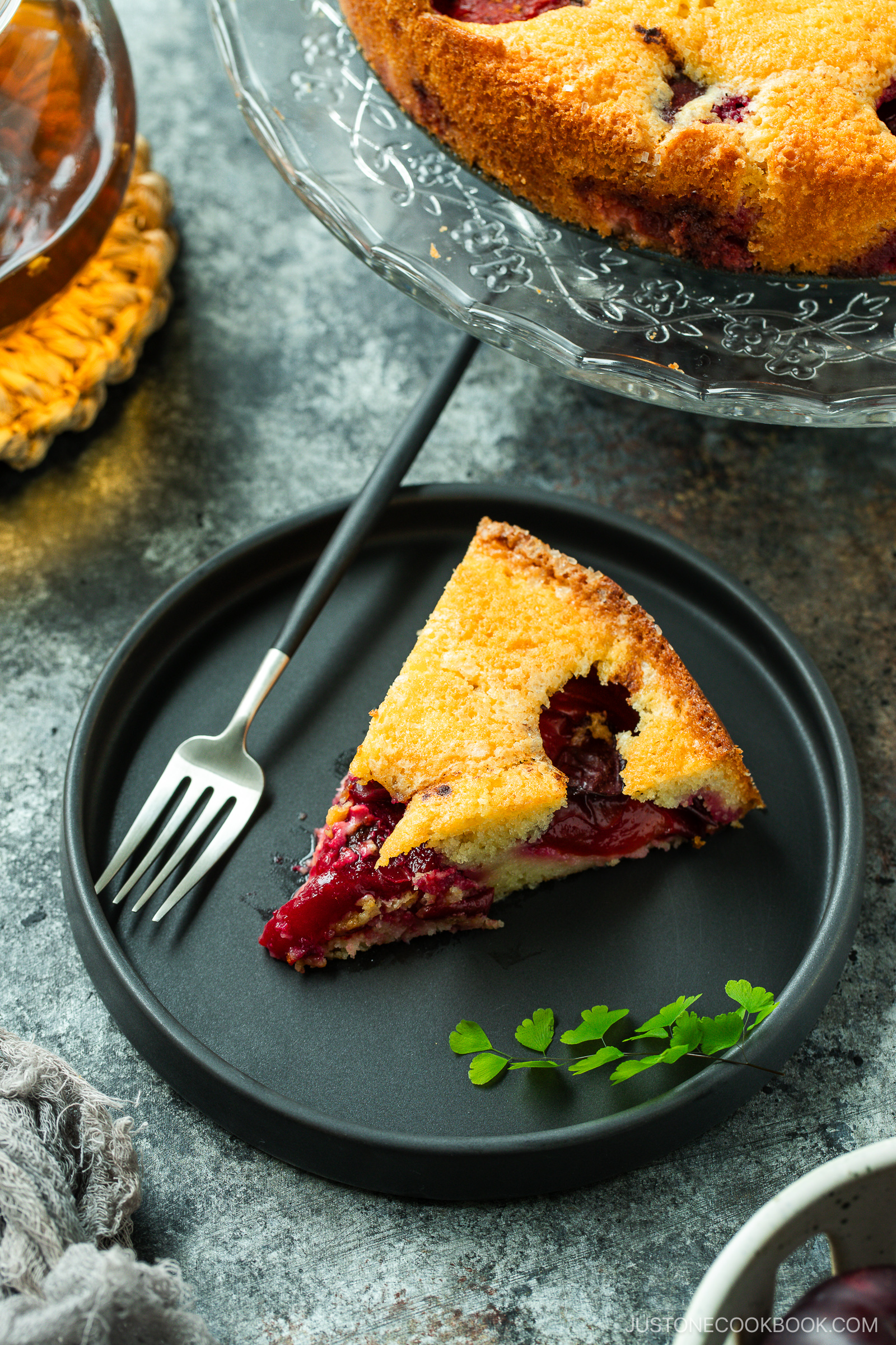 A slice of berry cake sits on a black plate with a fork beside it. The cake is golden brown with visible red berries, and a sprig of parsley is garnished on the plate. The rest of the cake is in the background on a glass stand.
